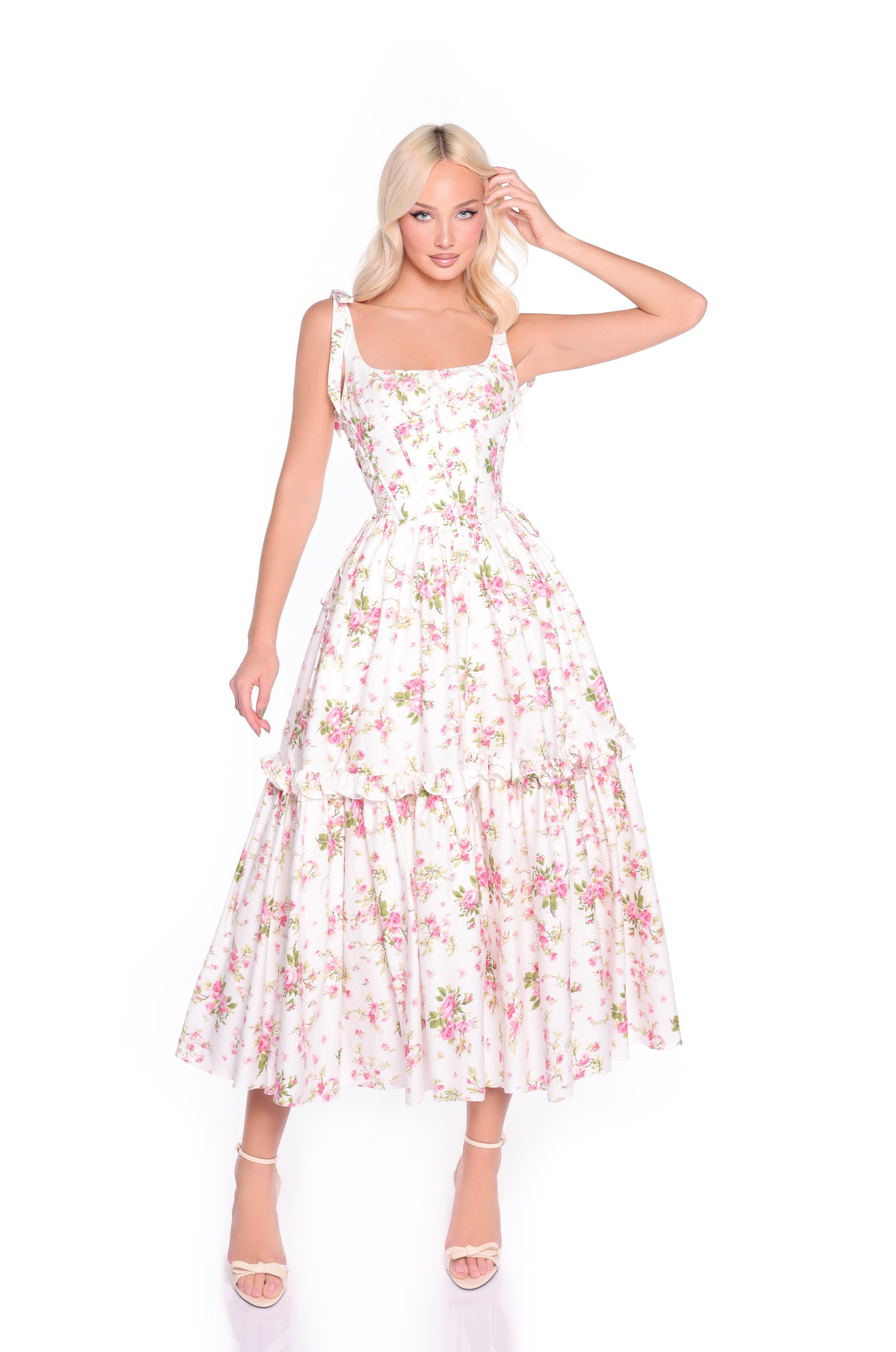 Woman wearing the Ivory Floral Casual Midi Corset Dress: Rosalie, a sleeveless fit-and-flare dress with pink and green floral print and ruffled tiers, paired with white high-heeled sandals, standing against a white background.